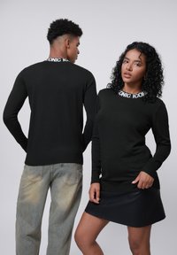 Zwarte trui met lange mouwen en geribbelde manchetten, met een witte "ICONIC"-logo op de nek. Deze is gecombineerd met licht vervaagde jeans en een zwarte leren minirok.