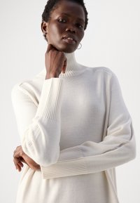 Pull à col roulé crème avec des détails côtelés au niveau du col et des poignets, présentant un tricot texturé et de subtiles perforations le long des manches.