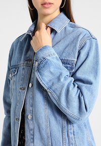 Chaqueta vaquera azul claro con un diseño clásico, que presenta un cierre de botones en la parte delantera, un gran cuello y detalles de costuras visibles en las mangas.