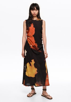Kobieta o prostych czarnych włosach, ubrana w czarną sukienkę bez rękawów z pomarańczowymi, żółtymi i beżowymi abstrakcyjnymi wzorami oraz brązowymi sandałami.