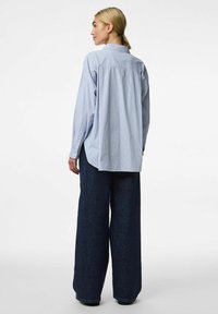 Camicia a righe blu chiaro e bianca, con vestibilità rilassata, maniche lunghe e dettaglio di piega sul retro. Abbinata a pantaloni in denim scuri a gamba larga.