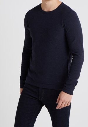 Mand iført en tætsiddende mørkeblå crewneck sweater og mørke jeans, stående mod en ensfarvet lys baggrund.