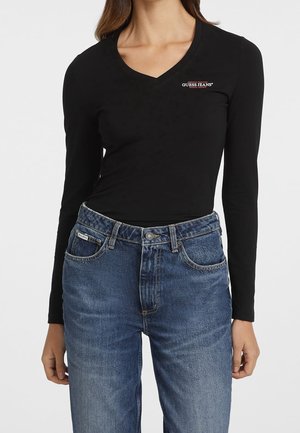 Kobieta ubrana w czarną, długą koszulkę z dekoltem w serek i logo Guess Jeans oraz niebieskie, wysokie dżinsy na jednolitym tle.