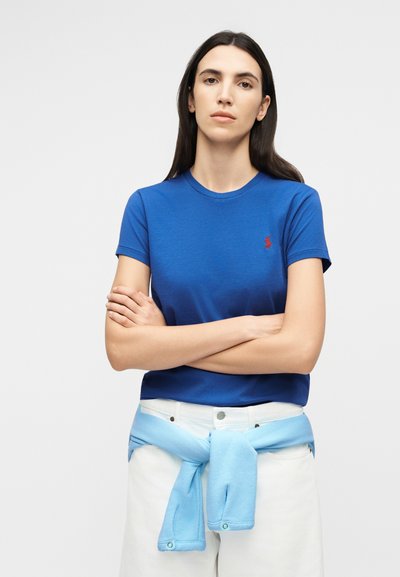 Γυναίκα με μακριά σκούρα μαλλιά που φοράει μπλε κοντομάνικο πουκάμισο και λευκό παντελόνι, με ένα ανοιχτό γαλάζιο πουλόβερ δεμένο γύρω από τη μέση, με σταυρωμένα τα χέρια.