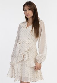 Rochie bej deschis cu mâneci lungi, decorată cu volane și talie strânsă. Confecționată dintr-un material transparent cu un model discret de puncte.