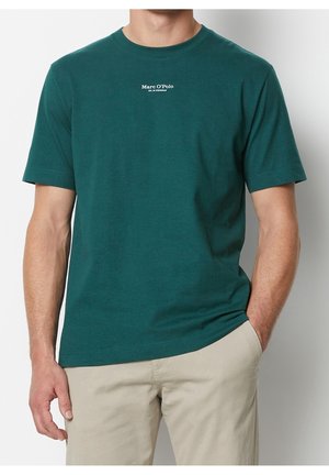 Mand iført en grøn kortærmet t-shirt med lille hvid tekst på brystet og beige bukser, den ene hånd i lommen, mod en ensfarvet baggrund.