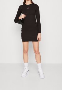 Black long-sleeve dress with round neckline and fitted silhouette. Features a small, multicolor logo on the chest. Model carries a black bag.