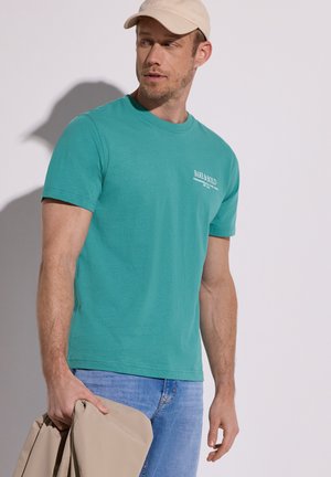 Homme portant un t-shirt bleu sarcelle et une casquette beige tient une veste beige, regardant sur le côté devant un fond clair uni.