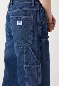 Un pantalon cargo en denim bleu foncé avec de multiples poches, un patch de marque à l'arrière et des coutures contrastées pour une texture supplémentaire.
