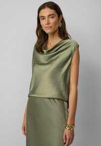 Cowlneck-top in olijfgroen satijn, zachte textuur, mouwloos ontwerp, gecombineerd met een bijpassende rok en een gestreepte armband om de pols.
