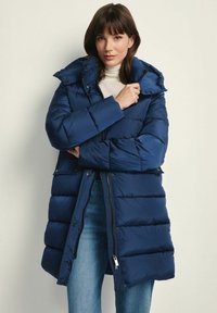 Granatowa kurtka puchowa z zamkiem z przodu, dużym kapturem i pikowanym wzorem. W zestawie z jasnym topem i dżinsowymi spodniami.