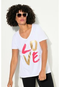 T-shirt blanc avec un audacieux graphisme "LOVE" en or et rose, offrant une coupe décontractée et des manches courtes. Le modèle porte des lunettes de soleil marron oversize.