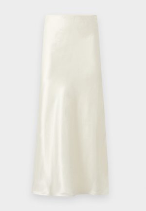 Femme aux longs cheveux foncés portant un corset crème sans bretelles et une longue jupe assortie, posant avec une main sur la hanche, fond neutre.