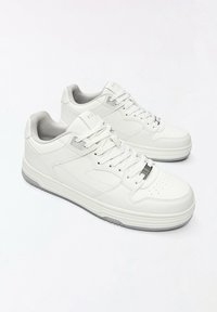 Paire de baskets basses blanches avec lacets, zone des orteils perforée et semelles gris clair sur fond blanc uni.