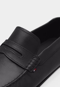 Μαύρο δερμάτινο λοφάρ με ραφές μοκασίνου και μικρή υφασμάτινη ετικέτα σε κόκκινο, λευκό και μπλε στο πλάι κοντά στη φτέρνα.