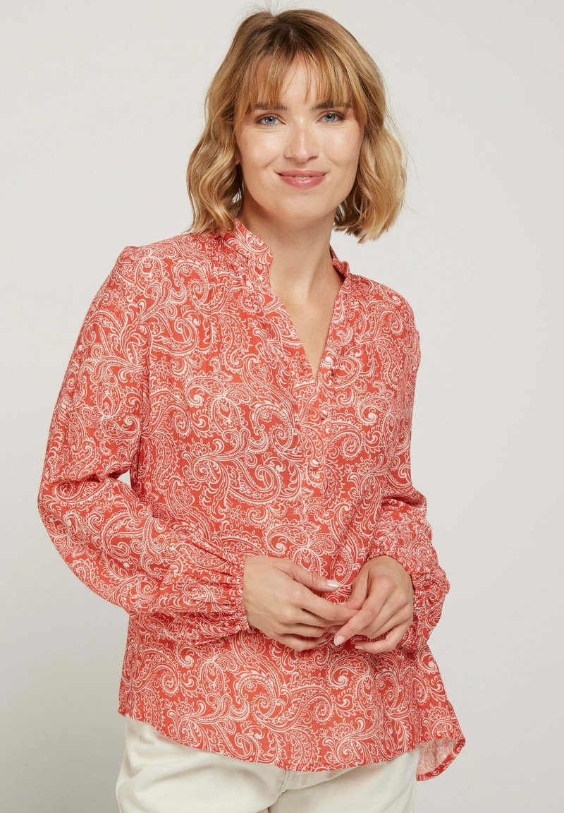 Blouse rouge à motif paisley avec un col montant, une patte de boutonnage et des manches froncées. Tissu lisse avec un léger éclat.
