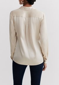 Beige Satinbluse mit langen Ärmeln, einem Kragen und subtilem Glanz. Verfügt über einen geraden Schnitt und knöpfbare Manschetten, kombiniert mit dunklen Jeans.