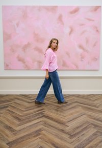 Femme en blouse rose et jean large marchant sur un sol en bois à chevrons devant une grande œuvre d'art murale abstraite rose et beige.