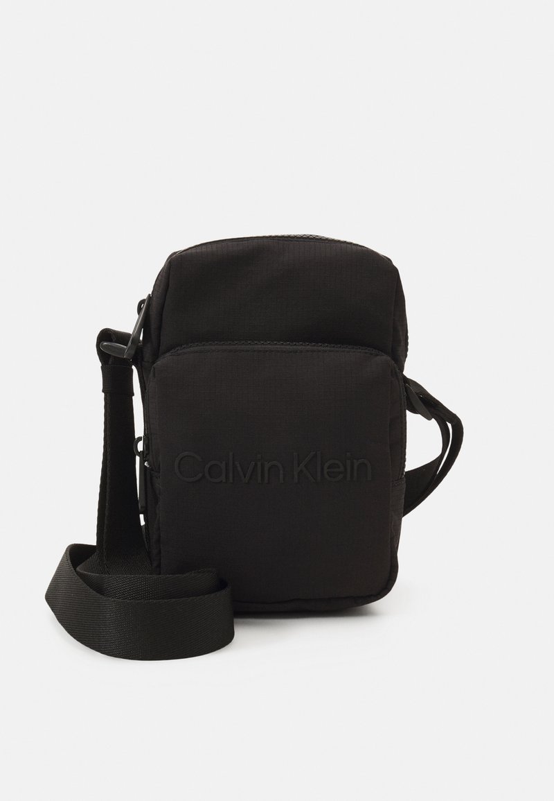 Borsa a tracolla rettangolare nera in tessuto testurizzato, con zip superiore, tracolla regolabile e logo "Calvin Klein" in rilievo sul fronte.