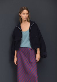 Īss tumši zils žakete ar rāvējslēdzēju priekšpusē un sānu kabatām, savienots ar gaiši zilu tanktopu un garu svītrainu sarkano un tumši zilo svārku.