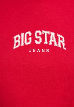Czerwona bawełniana koszulka z ribbowanym wzorem, na której białe litery w formie bloku układają się w napis "BIG STAR" nad "JEANS" w zakrzywionym układzie.