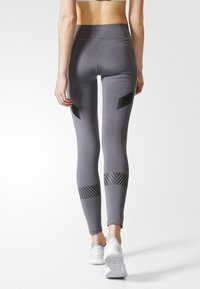 Šedé legíny s černými geometrickými akcenty, s vysokým páskem a texturovanými panely na dolní části nohou. Nosené s bílými teniskami.