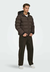 Giacca a vento marrone con cappuccio, caratterizzata da sezioni trapuntate; abbinata a jeans scuri e sneakers bianche, in piedi di fronte a uno sfondo chiaro.