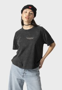 Camiseta negra de textura con mangas cortas, que presenta un gráfico "SINFONÍA VOLCÁNICA DE DESTRUCCIÓN" en el frente. Combinada con jeans de color azul claro.