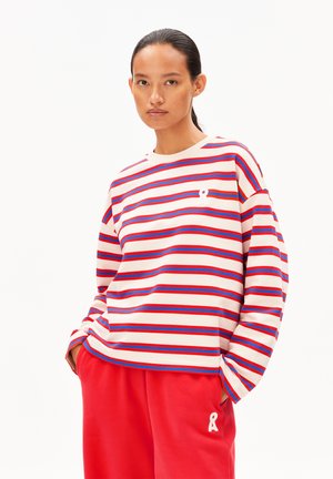 Vrouw met donker haar in een staart, gekleed in een crèmekleurig langmouw-shirt met rode en blauwe strepen en rode broeken, met haar handen in de zakken.