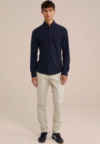 Cămașă albastră închisă cu mânecă lungă, cu nasturi, din țesătură, asociată cu pantaloni bej deschis și pantofi sport negri cu dungi albe.