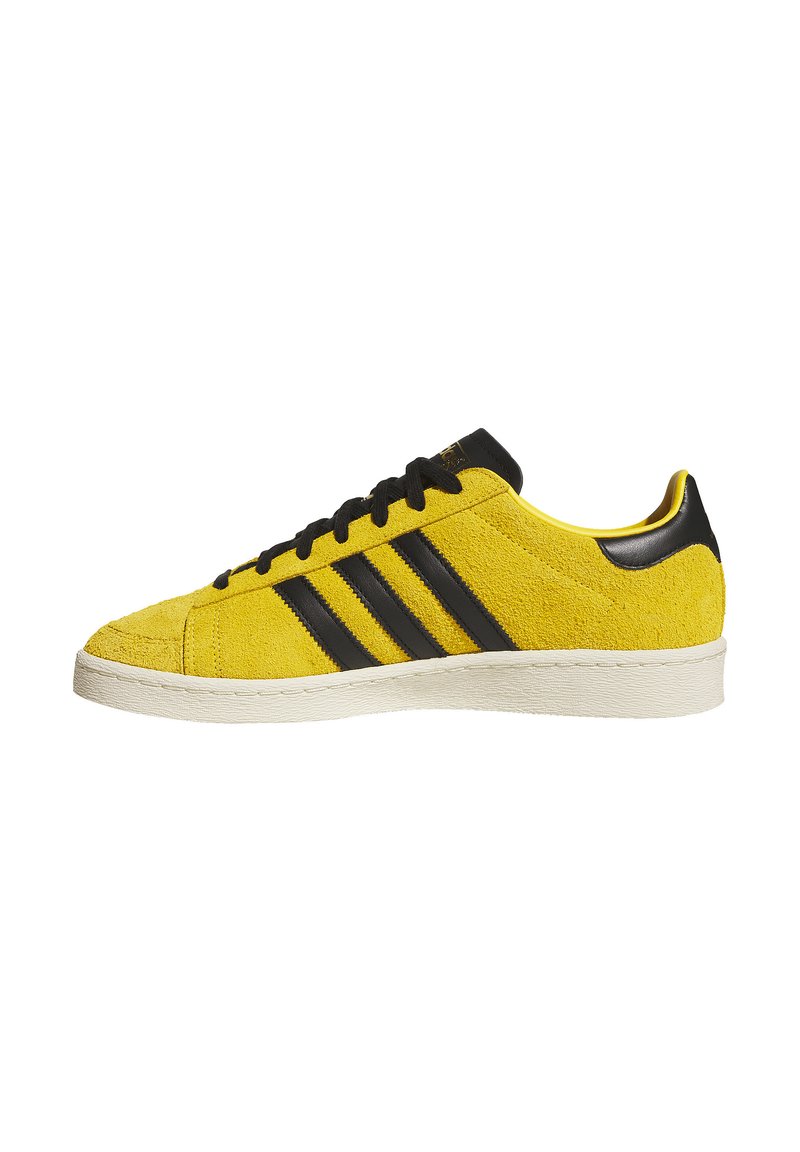 Gelber Wildledersneaker mit schwarzen Streifen, einer strukturierten Oberseite und einer weißen Gummisohle. Schwarze Akzente an Ferse und Zunge.