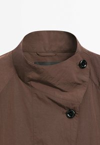 Bruine jas met een hoge kraag, voorzien van een gestructureerd oppervlak en een zwarte knoop. Rechte naden en een subtiel gestreept patroon zijn zichtbaar.