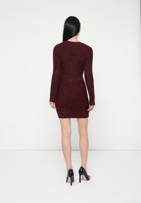 Abito lavorato a maglia bordeaux con un motivo testurizzato, maniche lunghe, vita e orlo a costine, abbinato a scarpe con tacco nere.