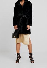 Sort faux fur frakke med bælte i taljen, lange ærmer og sjalkrave. Sammen med en silkeagtig guldskørt, sorte strophæle og en lille sort taske.