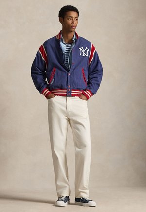 Ung mand iført en blå og rød varsityjakke, stribet skjorte, hvide bukser og marineblå sneakers, stående med hænderne i lommerne.