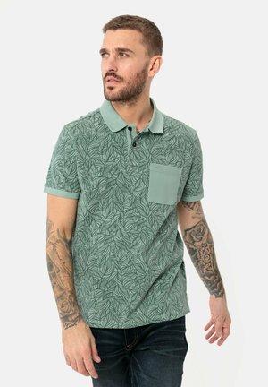 Groene polo shirt met korte mouwen en bladerenpatroon, geribbelde kraag, een drieknoopsrijg en borstzak. Draagt bij donkere spijkerbroek.