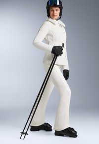 Giacca da sci bianca con trama a maglia a trecce, pantaloni da sci bianchi coordinati, guanti neri e bastoncini da sci; il modello indossa un casco nero e occhiali da sci.