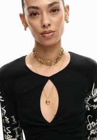 Zwart geribd top met lange mouwen en een keyhole uitgesneden detail, met witte handgeschreven tekst op de mouwen. Gouden ketting met hangeraccenten.