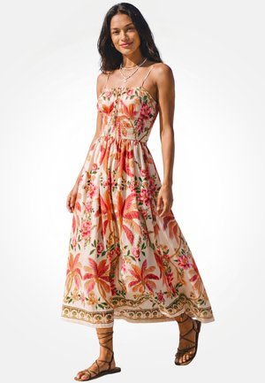 Donna che indossa un vestito lungo senza maniche con stampa floreale, spalline sottili e sandali piatti marroni con cinturini, in piedi su sfondo bianco.