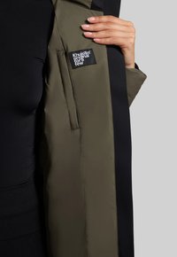 Bunda s kapucí v olivově zelené barvě, s černými akcenty a logem. Má hladkou strukturu a design bočních kapes.