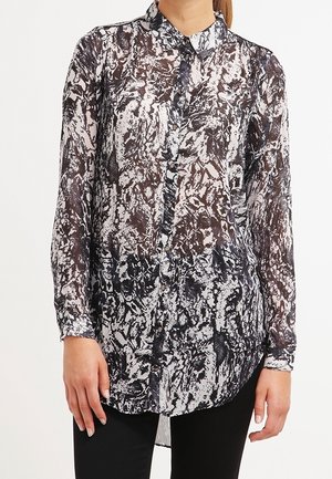 Kobieta w przezroczystej bluzce na guziki z długim rękawem w abstrakcyjny czarno-biały wzór, z kołnierzykiem, oraz w czarnych spodniach.