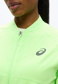 Εγγύς πλάνο ατόμου που φοράει φούξια ανοιχτό πράσινο μπουφάν με φερμουάρ, με διάτρητες λεπτομέρειες και μαύρο αθλητικό λογότυπο στο στήθος.