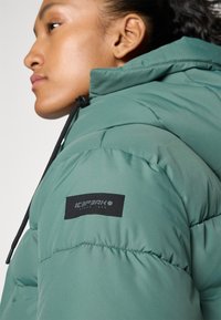 Kvinna som bär en grön puffjacka med huva, som har en svart lapplabel på överarmen där det står "ICEPEAK."