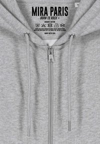 Felpa con zip in cotone organico grigio, dotata di coulisse, con una zip YKK e un'etichetta che riporta "MIRA PARIS—BORN TO ROCK."