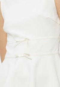 Primo piano di un vestito bianco senza maniche con due sottili legacci orizzontali intorno alla vita su una persona dalla pelle chiara.