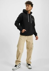 Svart hettegenser med frontlomme, hvite snorer, kombinert med beige cargobukser. Modellen har på seg svart-hvite joggesko og står foran en nøytral bakgrunn.
