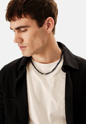 Náhrdelník z korálků s kulatými tmavě modrými a černými kameny, nošený přes bílou košili a černou bundu; hladká textura a jemný lesk.