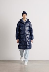Geacă puffer bleumarin cu glugă, cu un design matlasat, butoni frontali și buzunare laterale. Purtați peste o cămașă ușoară și blugi casual.
