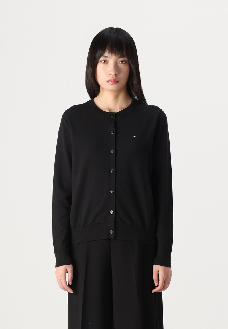 Zwarte cardigan van zacht materiaal, met een ronde halslijn, knoopsluiting en een klein logo-detail. Lange mouwen met ribgebreide boorden.