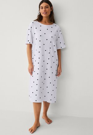 Vrouw staat blootsvoets, draagt een witte jurk tot op de knie met korte mouwen en zwarte hartjespatronen, glimlacht lichtjes.